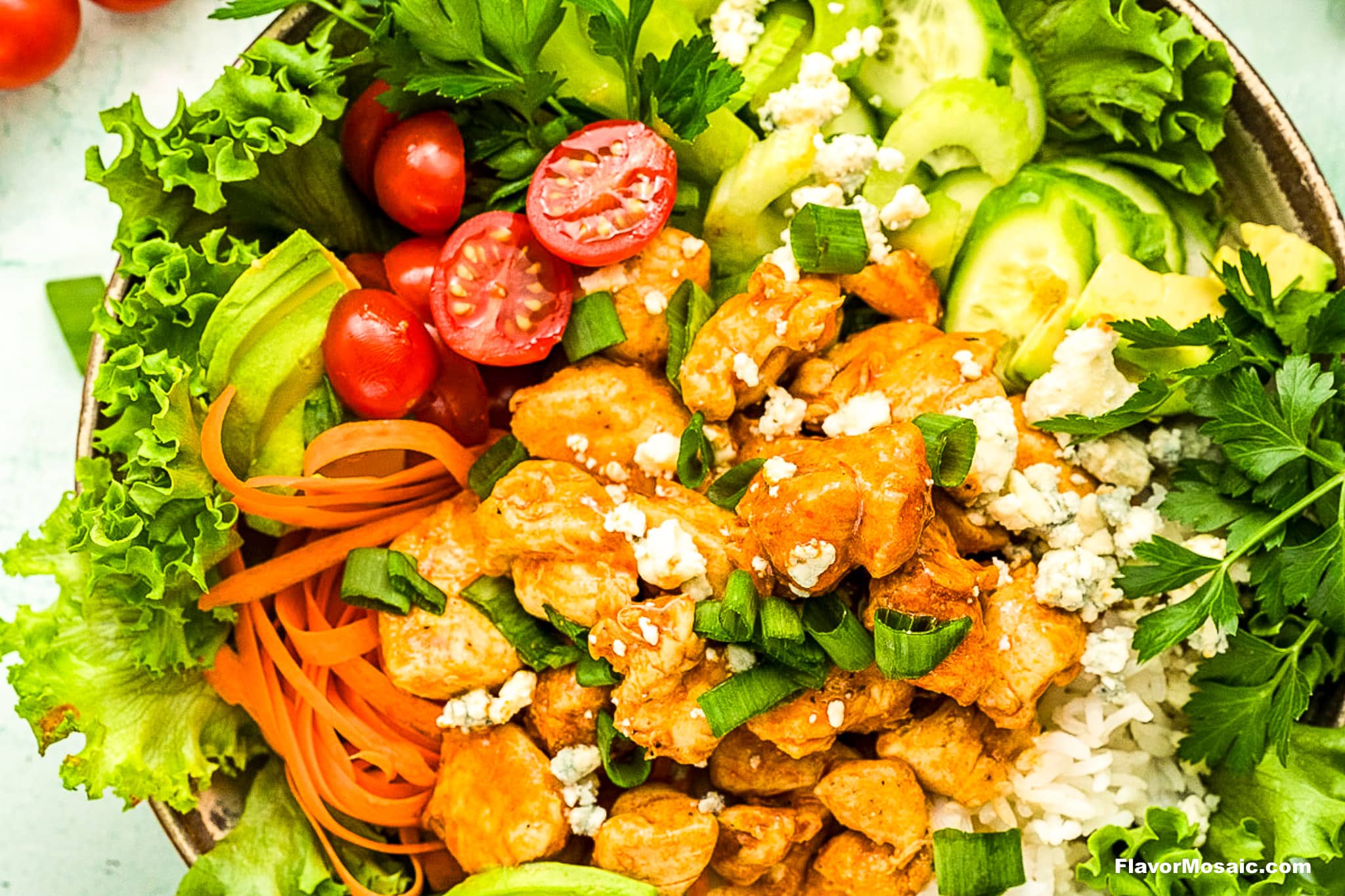A colorful bowl filled with lettuce, cherry tomatoes, cucumber, sliced avocado, shredded carrots, rice, and seasoned chicken topped with blue cheese and chopped green onions. Fresh parsley garnishes the bowl.