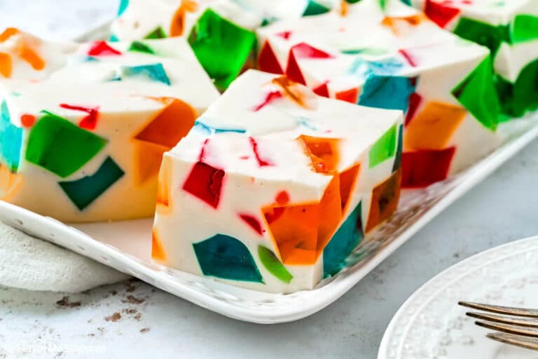 Colorful mosaic gelatin dessert squares with chunks of red, green, orange, and blue gelatin set in a creamy white base, arranged on a white serving plate. A fork and part of a plate are visible nearby.