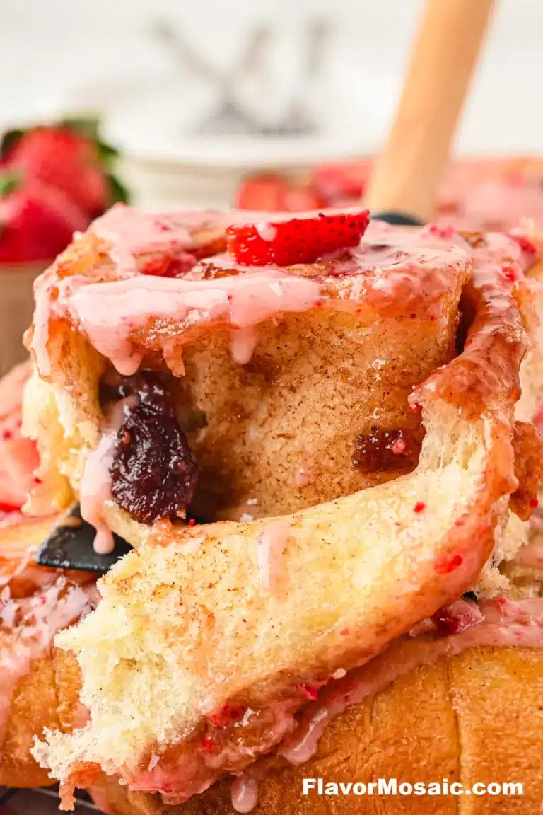 A close-up of a gooey cinnamon roll topped with pink strawberry glaze and a slice of fresh strawberry, highlighting the ultimate in Strawberry Desserts, with more rolls and strawberries blurred in the background.