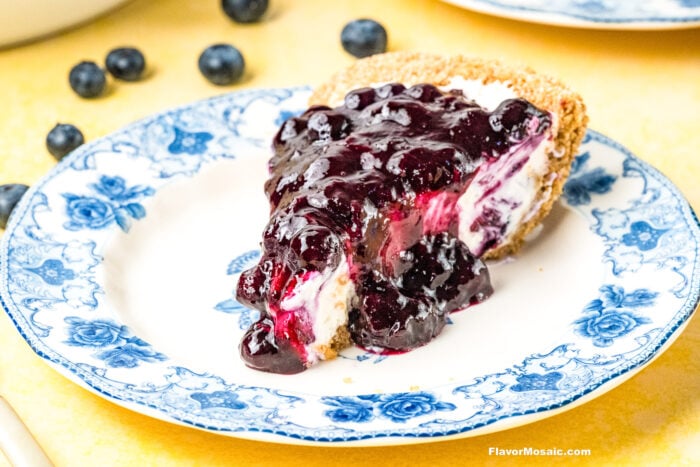 A slice of blueberry cheesecake with blueberry topping sits on a blue and white floral-patterned plate, surrounded by a few fresh blueberries on a yellow surface.
