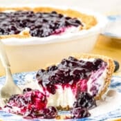 A slice of creamy cheesecake with a graham cracker crust and blueberry topping sits on a blue and white plate with a fork, in front of the remaining pie in a white dish.