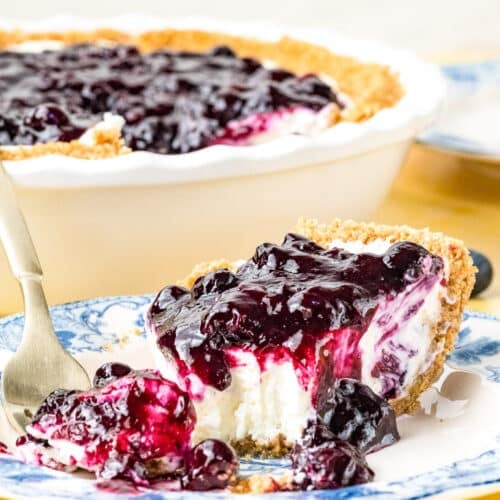 A slice of creamy cheesecake with a graham cracker crust and blueberry topping sits on a blue and white plate with a fork, in front of the remaining pie in a white dish.