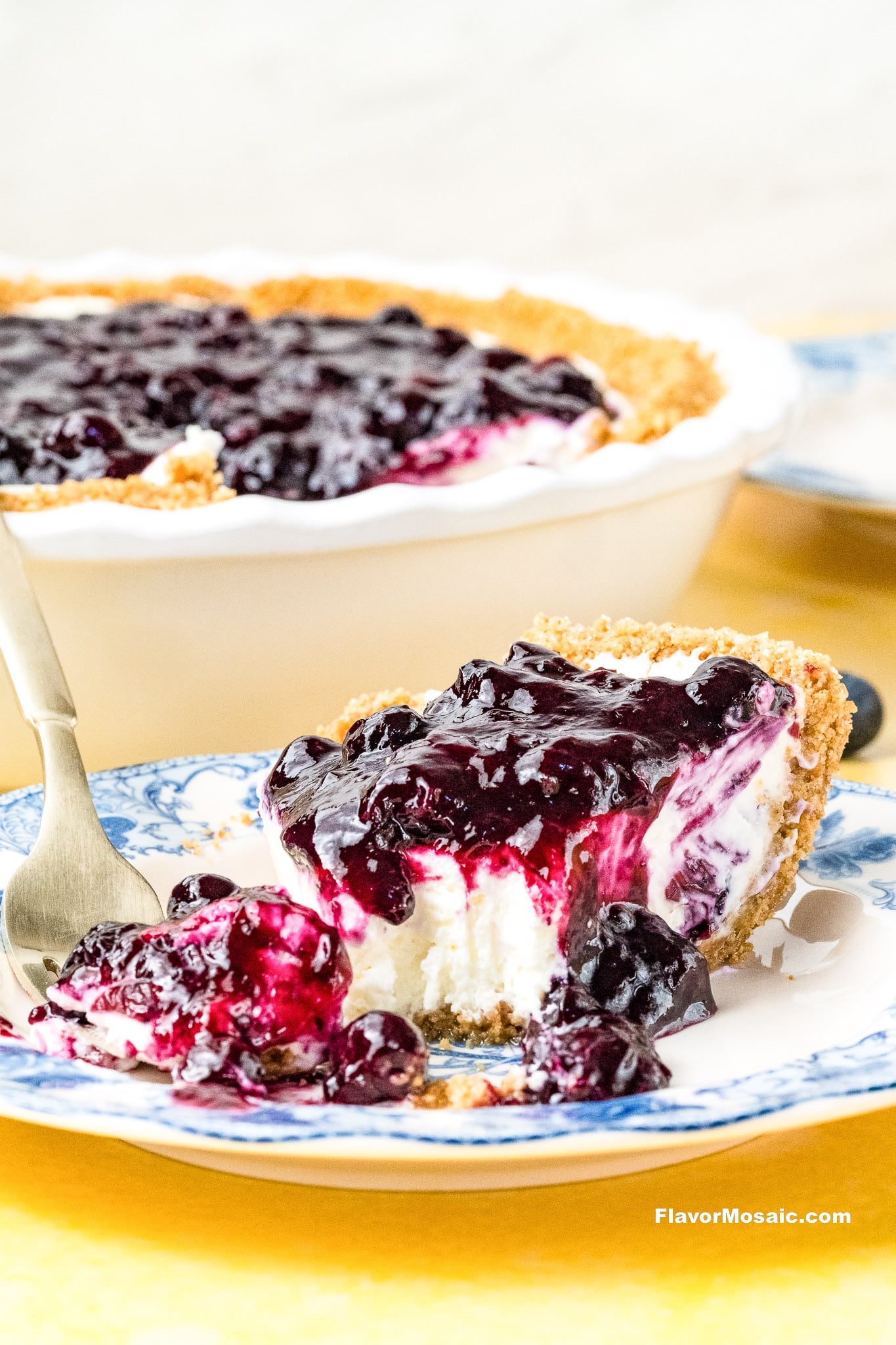 A slice of creamy cheesecake with a graham cracker crust and blueberry topping sits on a blue and white plate with a fork, in front of the remaining pie in a white dish.