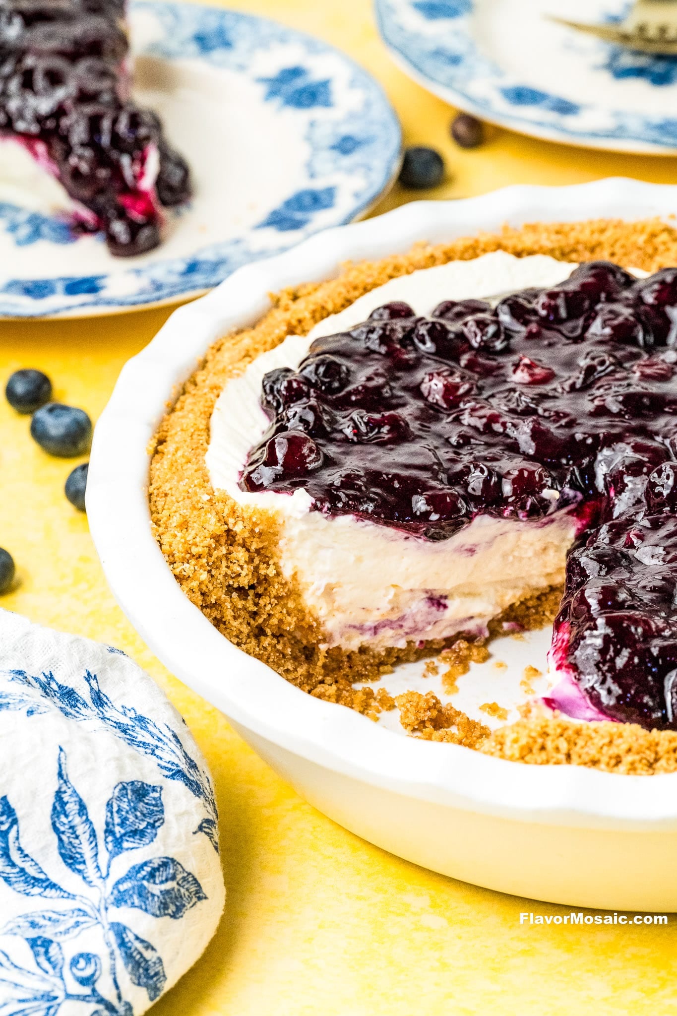 A cheesecake with a graham cracker crust topped with blueberry compote, missing a slice. The pie dish sits on a yellow surface, with blueberries and blue-patterned plates in the background.