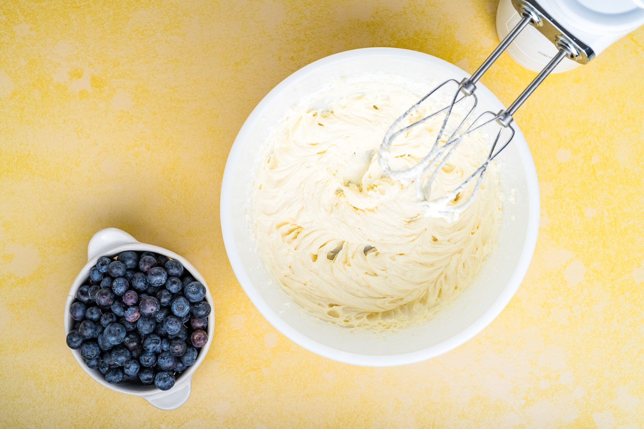 A white bowl of creamy batter-perfect for Blueberry Cream Cheese Pie-is being mixed with an electric hand mixer, sitting on a yellow surface next to a smaller bowl filled with fresh blueberries.