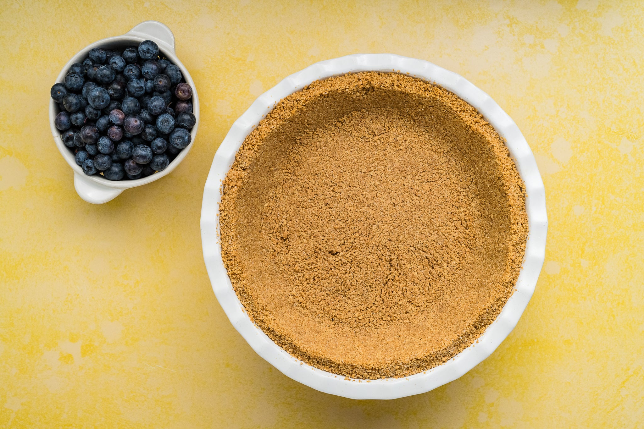 A white dish with a graham cracker pie crust, perfect for a Blueberry Cream Cheese Pie, sits next to a small white bowl of fresh blueberries on a yellow surface.
