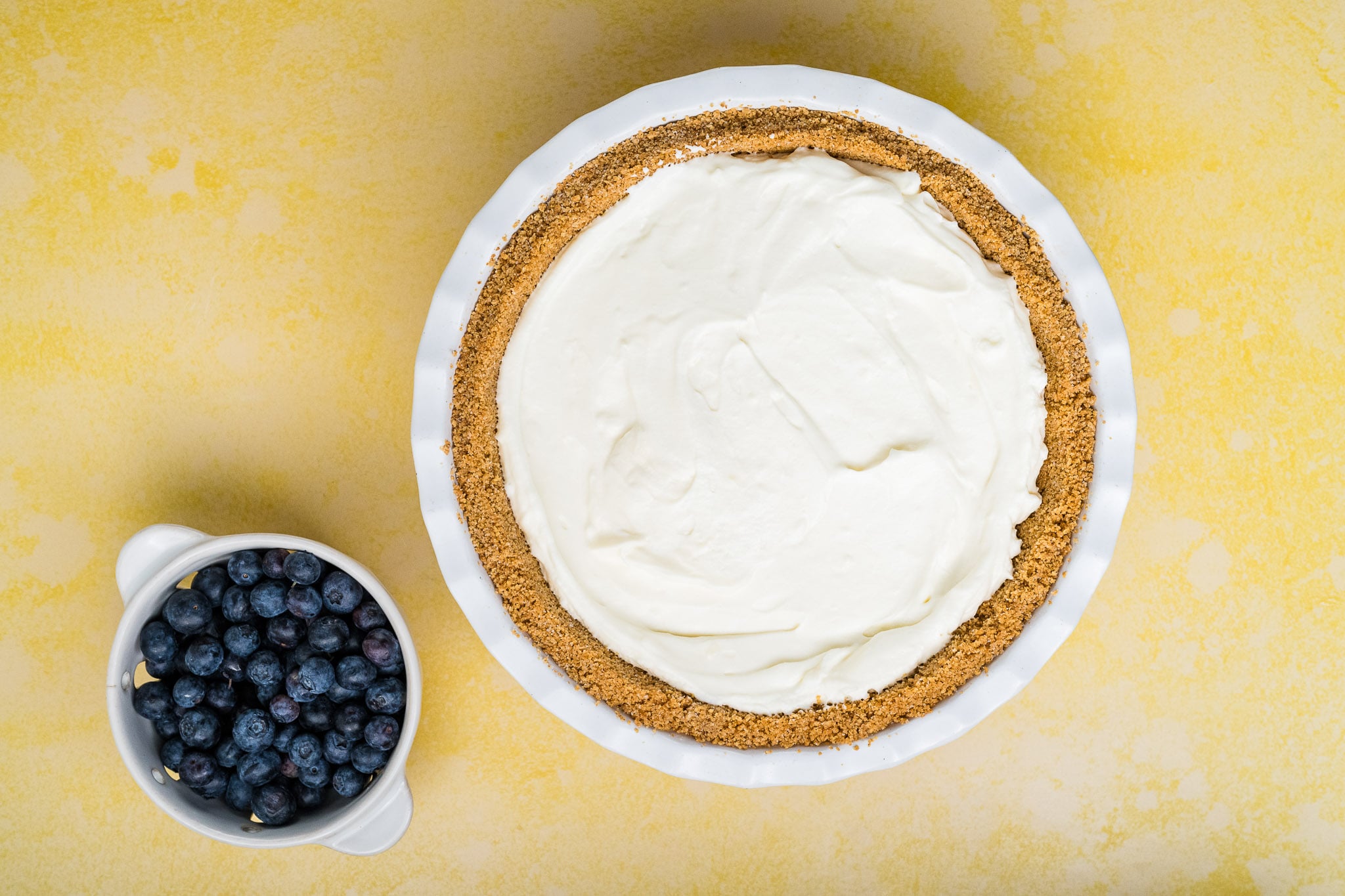 A Blueberry Cream Cheese Pie with a graham cracker crust and creamy white filling sits next to a small white pitcher filled with fresh blueberries on a yellow surface.