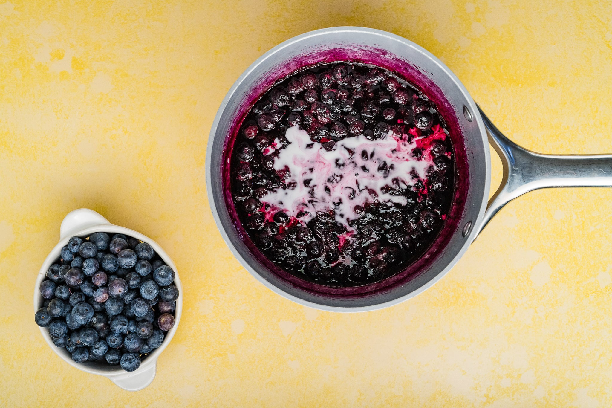A saucepan filled with cooked blueberries and a swirl of cream sits on a yellow surface next to a small white bowl of fresh blueberries-perfect ingredients for a delectable Blueberry Cream Cheese Pie.