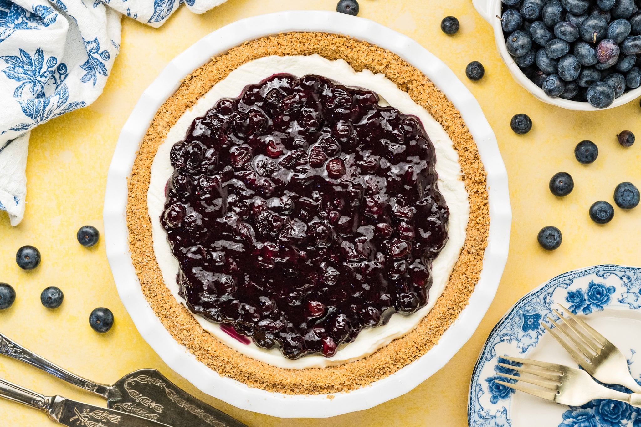 A Blueberry Cream Cheese Pie with a graham cracker crust and blueberry topping sits on a yellow surface, surrounded by a bowl of fresh blueberries, a blue-patterned plate with forks, and vintage serving utensils.