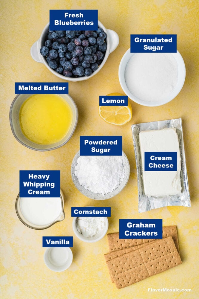 Top view of labeled ingredients for cheesecake: fresh blueberries, granulated sugar, melted butter, lemon, powdered sugar, cream cheese, heavy whipping cream, cornstarch, vanilla, and graham crackers on a yellow background.