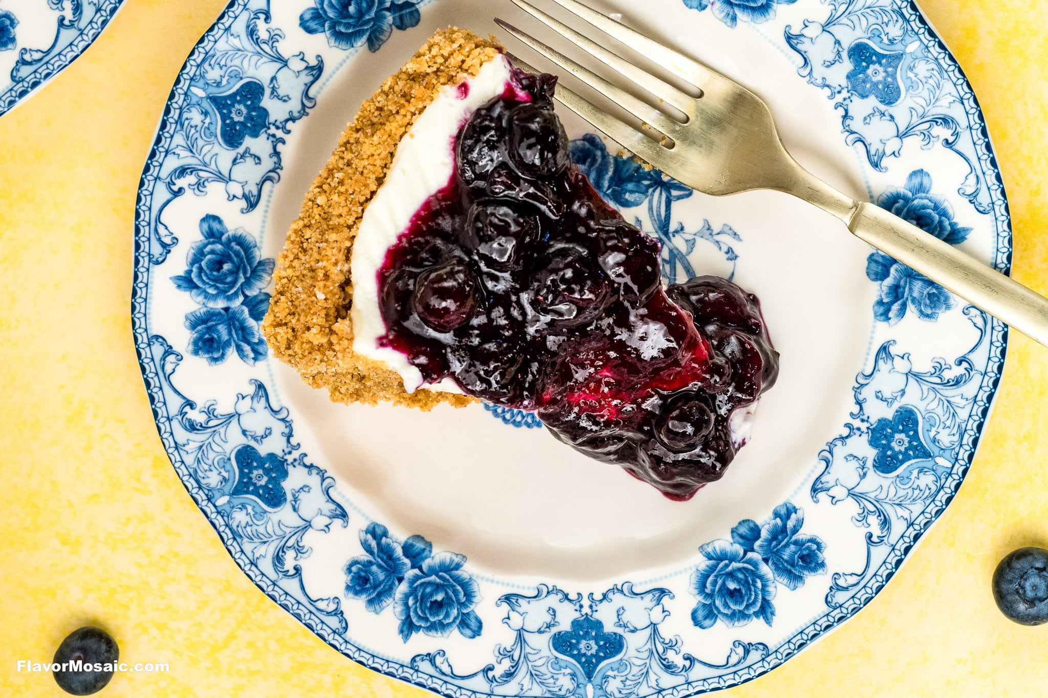 A slice of Blueberry Cream Cheese Pie with crumb crust and creamy filling sits on a white plate with blue floral patterns, beside a silver fork, set on a yellow surface.