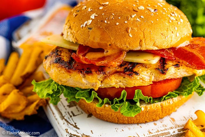 A grilled chicken sandwich with bacon, cheese, lettuce, and tomato on a sesame bun sits on a white wooden surface, with crinkle-cut fries visible in the background.