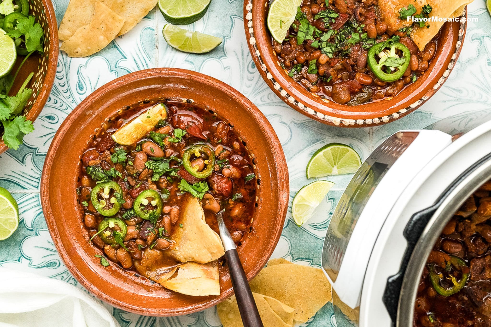 Two bowls of spicy pinto beans garnished with jalapeño slices, cilantro, lime wedges, and tortilla chips sit on a patterned surface alongside an Instant Pot, extra lime slices, cilantro, and tortilla chips.