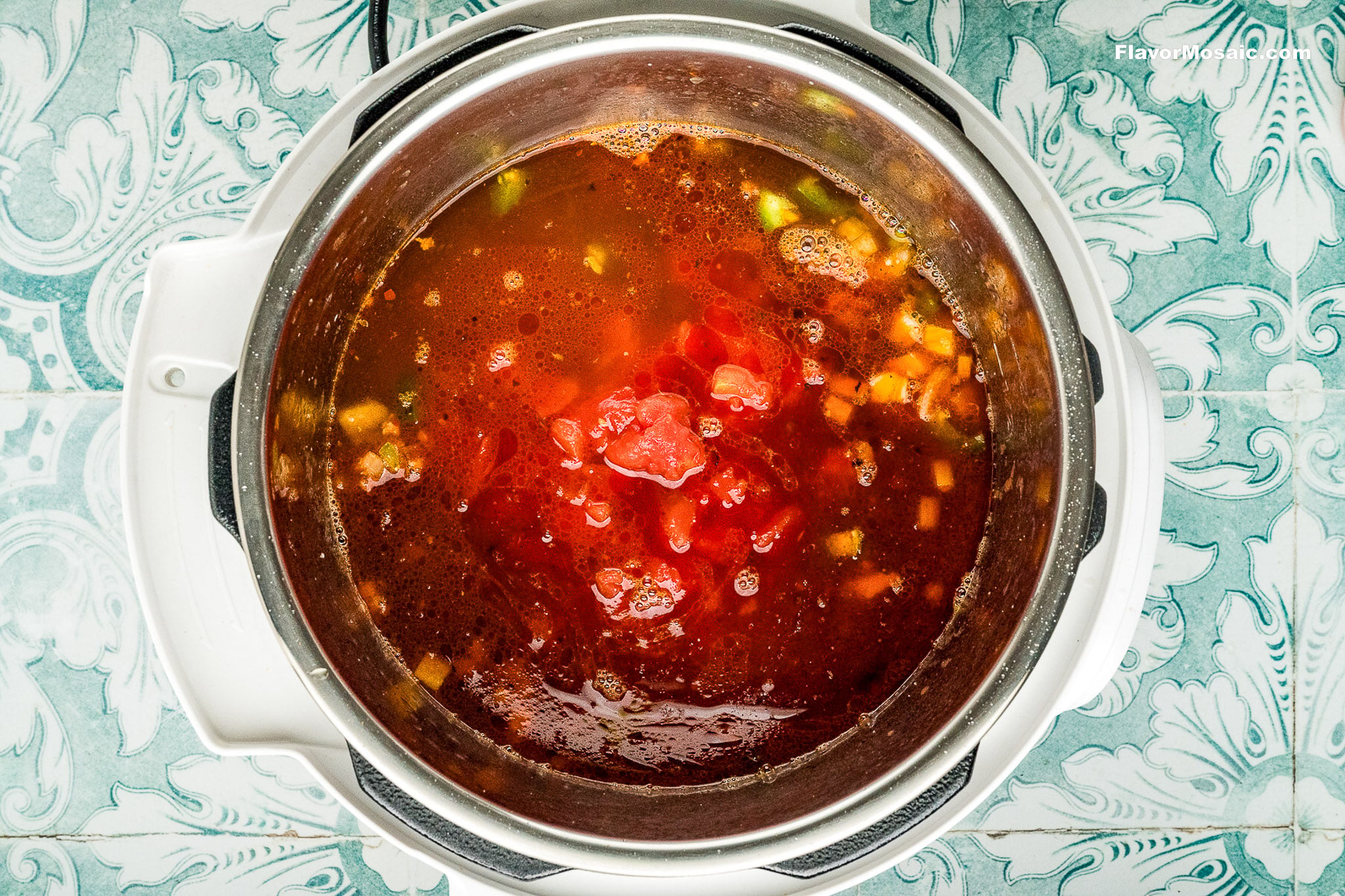 A top-down view of a pot filled with tomato-based soup, featuring chunks of tomatoes, vegetables, and broth-similar in style to Instant Pot Charro Beans (Frijoles Charros)-sitting on a patterned blue and white surface.