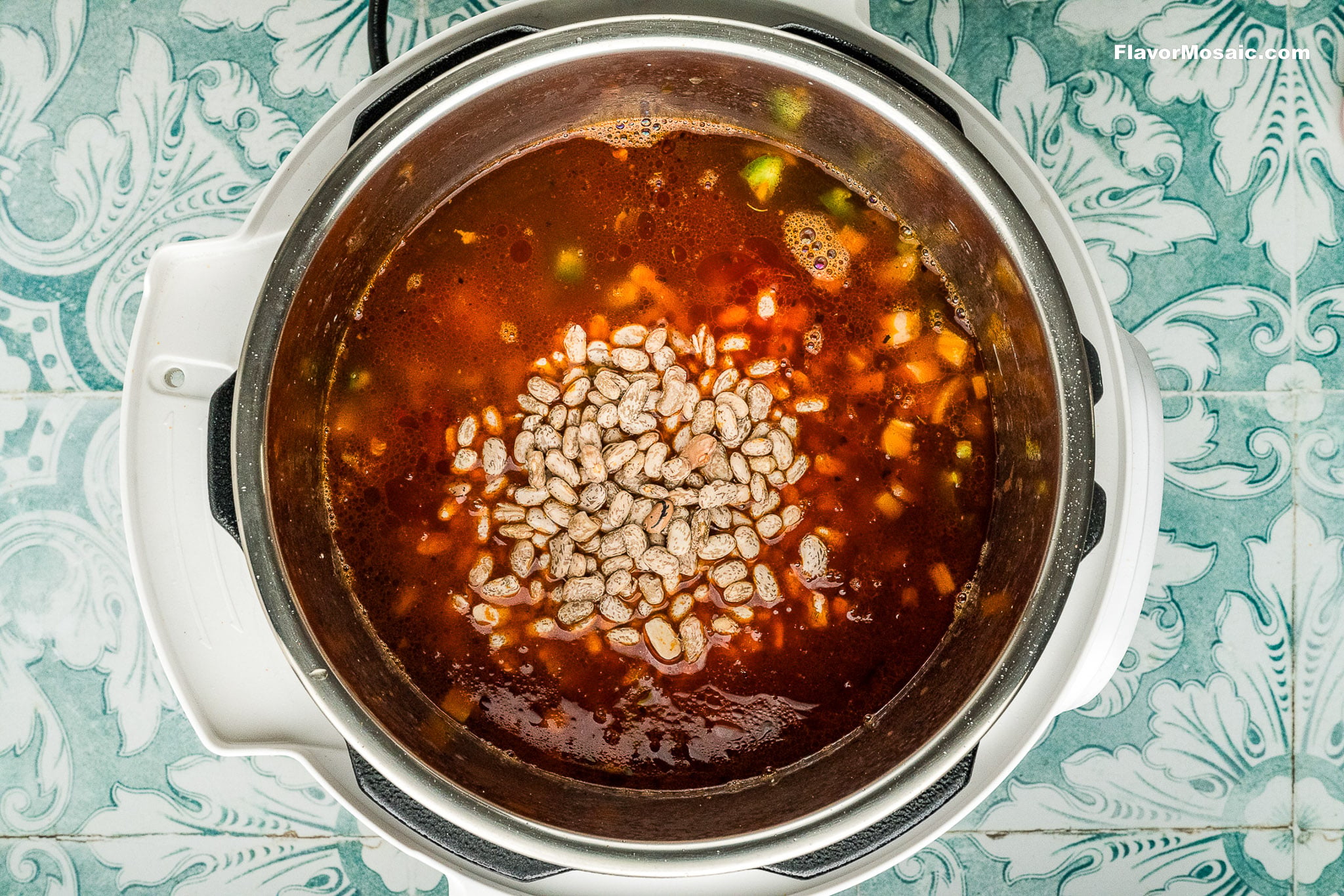 Overhead view of an Instant Pot filled with a red soup or stew for Instant Pot Charro Beans (Frijoles Charros), topped with uncooked pinto beans and chopped vegetables, set on a blue patterned surface.