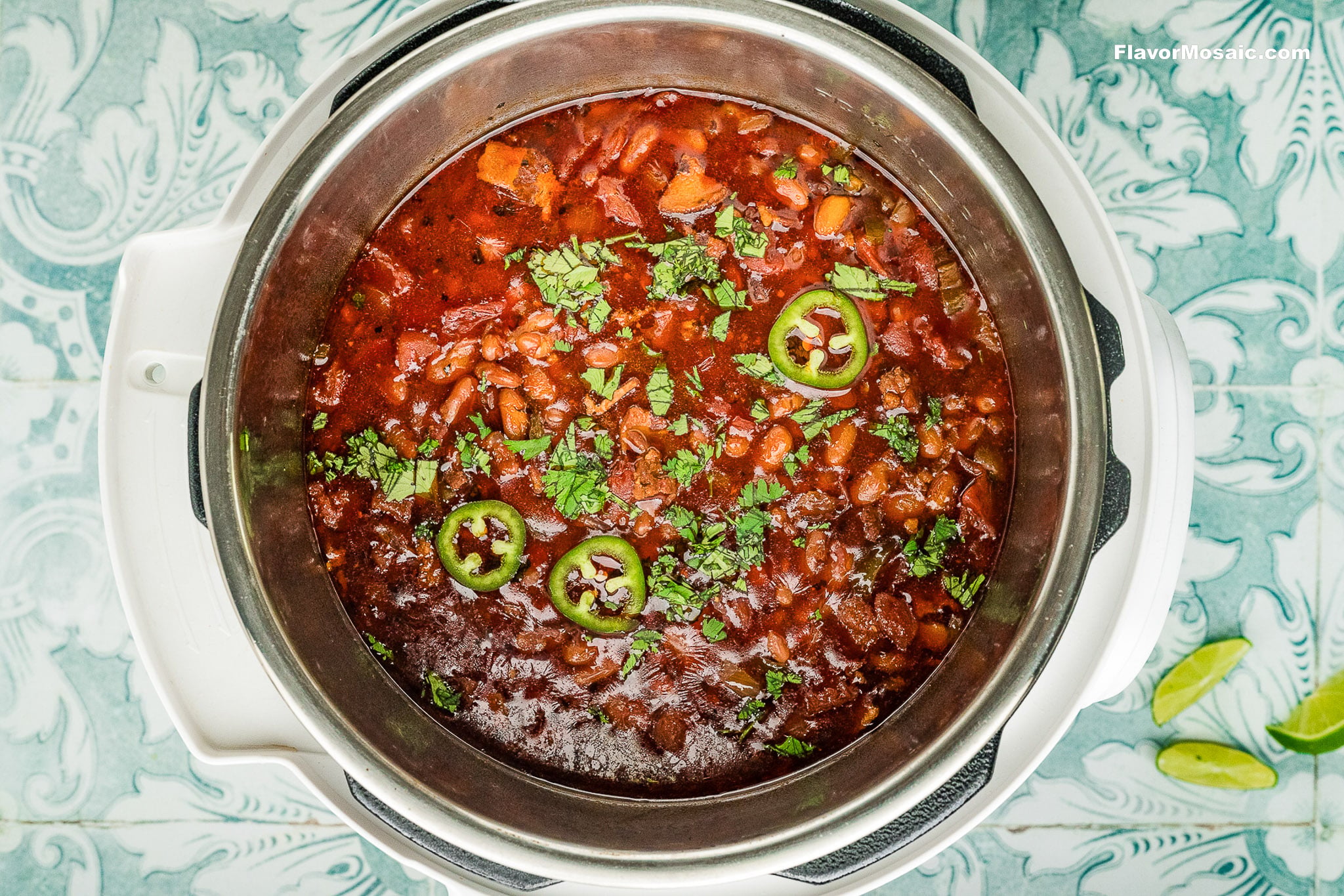 A pot of chili garnished with sliced jalapeños and chopped cilantro sits on a patterned surface with lime wedges nearby. The rich, hearty chili features beans and vegetables inspired by Instant Pot Charro Beans (Frijoles Charros).