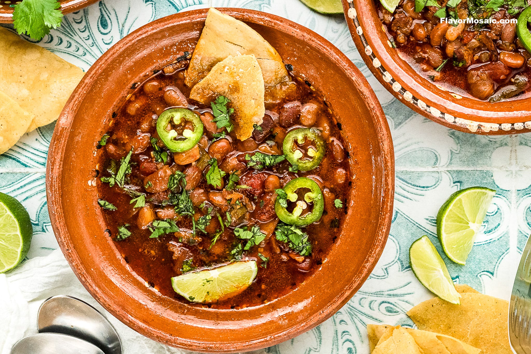 A bowl of chili garnished with jalapeño slices, chopped cilantro, tortilla chips, and lime wedges sits on a patterned surface. Nearby are extra limes, chips, and another bowl featuring hearty Instant Pot Charro Beans (Frijoles Charros).