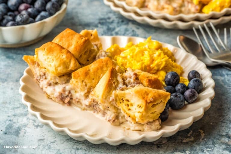 A plate with a biscuit-topped sausage gravy casserole, scrambled eggs, and fresh blueberries, with a fork and spoon beside it. In the background, another plate and a bowl of blueberries are visible.