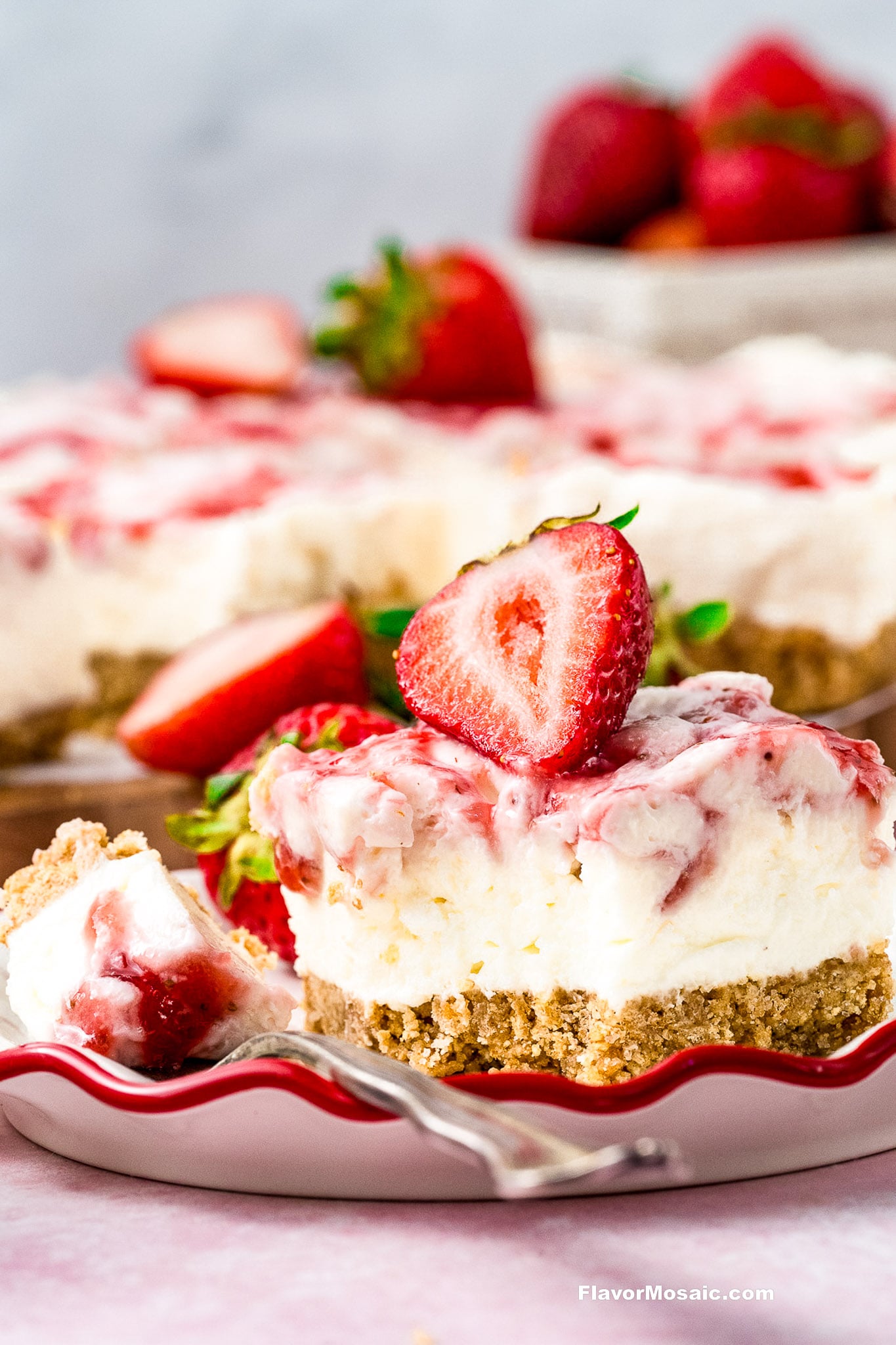 A slice of strawberry cheesecake with a crumbly crust is topped with a halved fresh strawberry. More cheesecake and whole strawberries are visible in the background, and a fork rests on the plate.
