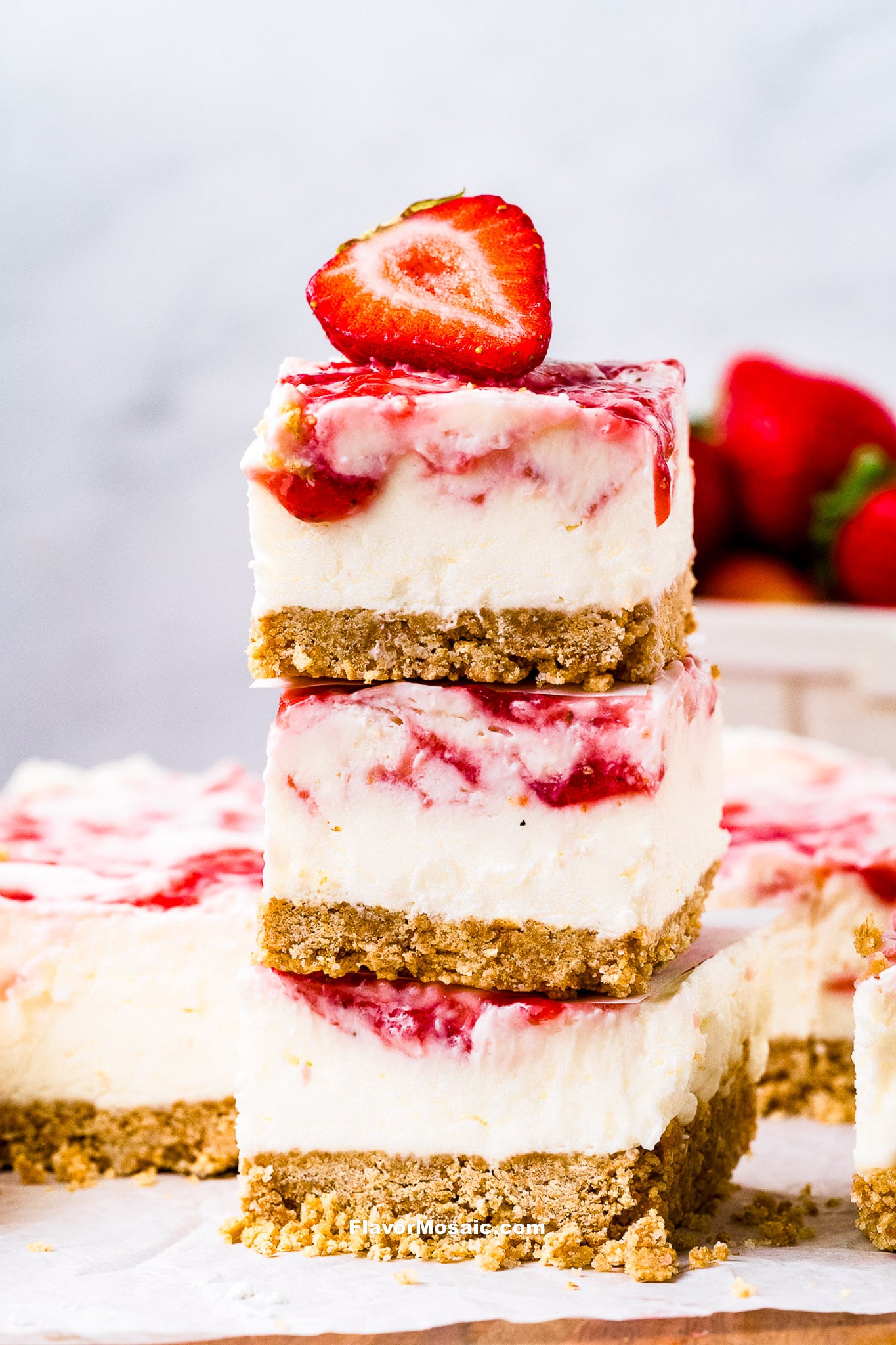 Three creamy strawberry cheesecake bars are stacked on top of each other, topped with a halved fresh strawberry. The bars have a crumbly cookie crust and swirls of strawberry sauce. Fresh strawberries are blurred in the background.