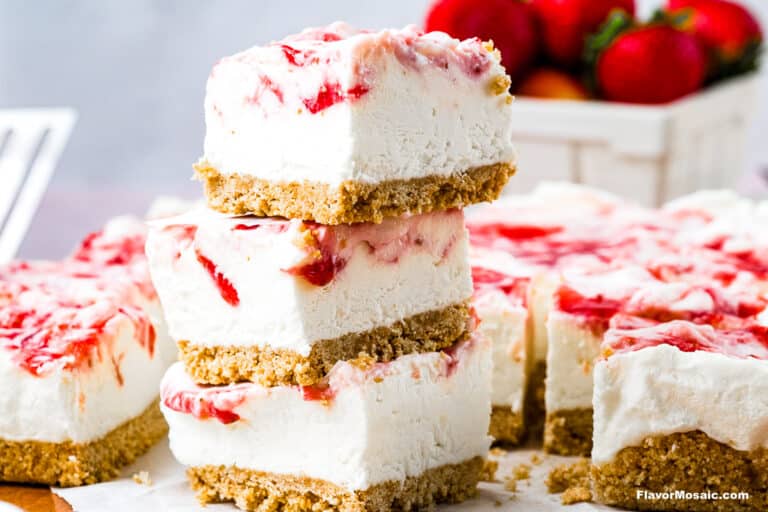 Three cheesecake bars with a graham cracker crust and swirls of strawberry sauce are stacked on top of each other, with more cheesecake bars and a basket of fresh strawberries in the background.