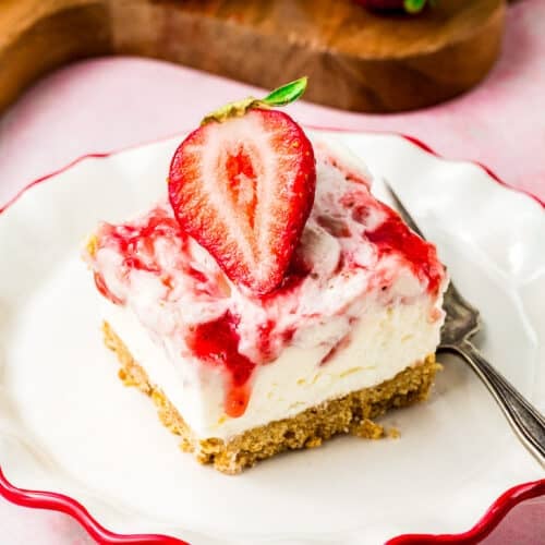 A strawberry cheesecake bar topped with a halved strawberry and strawberry sauce sits on a white plate with a red rim and a fork. A wooden board with more bars and strawberries is in the background.