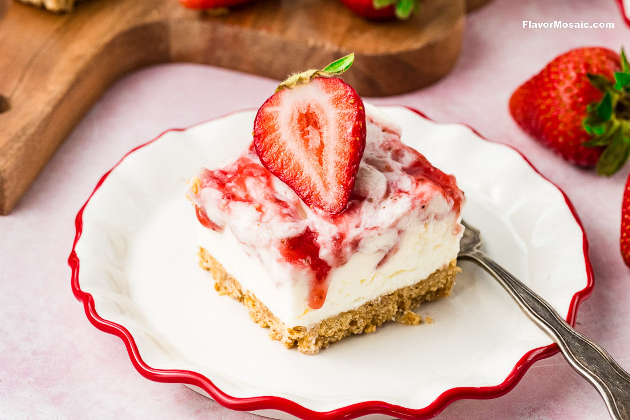 A slice of strawberry cheesecake with a graham cracker crust, topped with strawberry sauce and a halved strawberry, evokes the charm of Strawberry Cheesecake Bars, served on a white plate with fresh strawberries in the background.