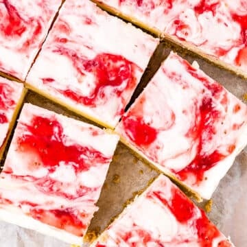 Nine Strawberry Cheesecake Bars rest on parchment paper, their creamy white and pink marbled tops adorned with vibrant red strawberry swirls, all perched on a golden brown crust.