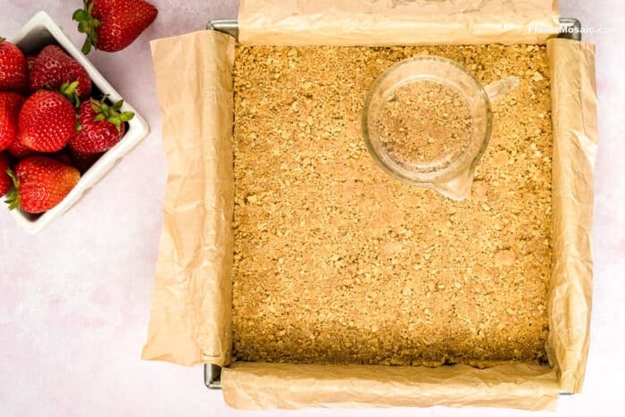A parchment-lined square baking pan filled with a pressed graham cracker crust, perfect for making Strawberry Cheesecake Bars. A small glass measuring cup sits on top, and a bowl of fresh strawberries is on the side.