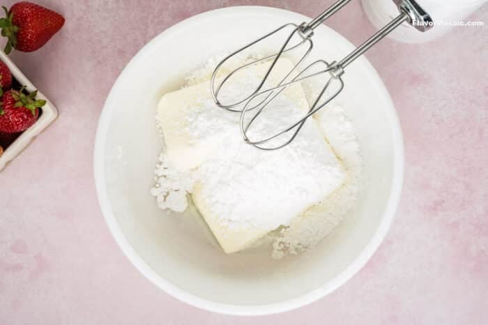 A white bowl with butter and powdered sugar, ready to be mixed with an electric hand mixer for Strawberry Cheesecake Bars; fresh strawberries are visible in a container next to the bowl on a pink surface.