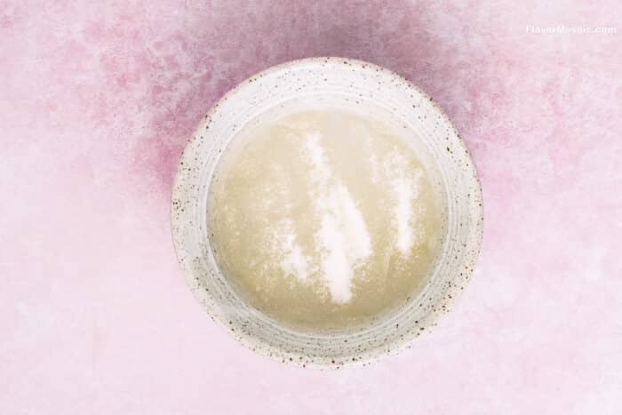 A speckled white bowl with a cloudy, pale liquid sits on a pink textured surface-perfect for mixing ingredients for Strawberry Cheesecake Bars. The liquid has streaks of white powder visible on top.