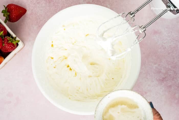 A hand holds a bowl of liquid over a large bowl of whipped cream, with an electric hand mixer and a container of strawberries nearby on a pink surface-perfect for making Strawberry Cheesecake Bars.