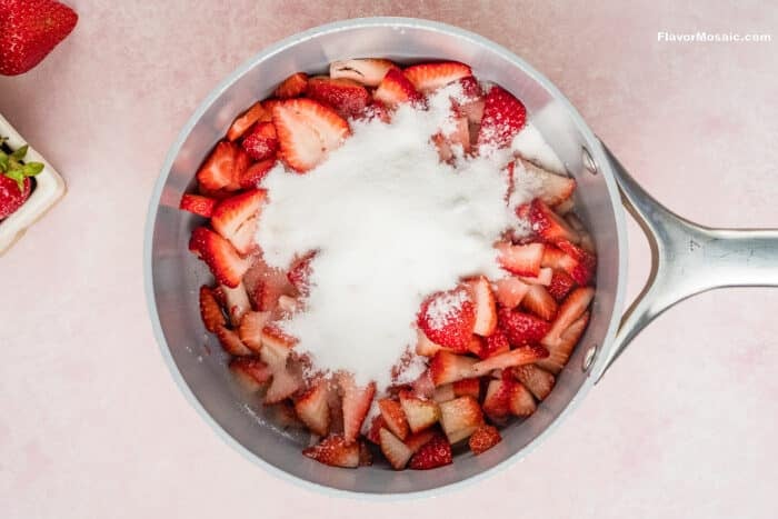 Overhead view of a saucepan filled with sliced strawberries and a mound of white sugar on top, perfect for making Strawberry Cheesecake Bars, on a light pink surface. Part of a plate with more strawberries is visible in the corner.
