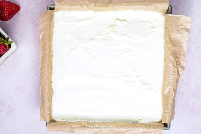 A square baking pan lined with parchment paper, filled with smooth, creamy Strawberry Cheesecake Bars. Part of a bowl of fresh strawberries is visible in the corner on a light pink surface.