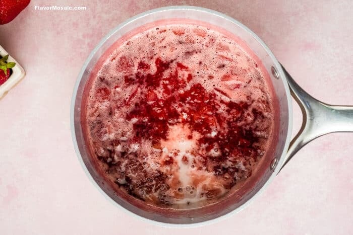 A saucepan filled with bubbling, partially mashed strawberries in a red syrup-perfect for topping Strawberry Cheesecake Bars-sits on a light pink surface, with some foam and chunks visible.