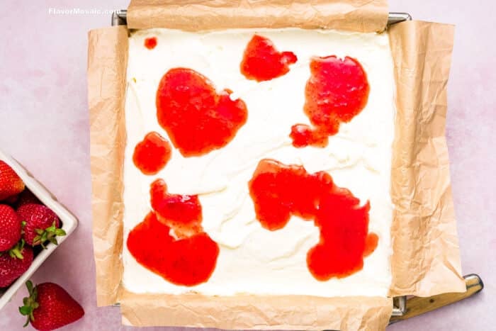 A square baking pan lined with parchment paper is filled with a creamy white mixture for Strawberry Cheesecake Bars, topped with large red strawberry sauce spots. A bowl of fresh strawberries sits beside the pan on a light pink surface.