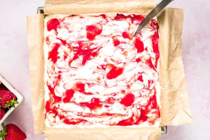 A square baking pan lined with parchment paper holds a swirled mixture of creamy white and bright red, resembling freshly made Strawberry Cheesecake Bars. A spoon rests in the pan, with fresh strawberries nearby.