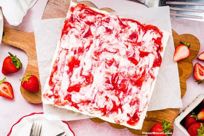 A square Strawberry Cheesecake Bar with a swirled pattern of red strawberry sauce and creamy white filling sits on parchment paper, surrounded by fresh strawberries and baking tools.
