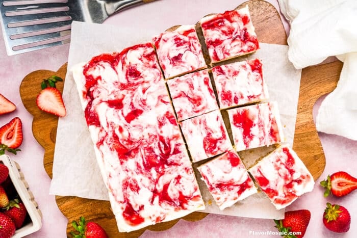 A batch of Strawberry Cheesecake Bars cut into squares on parchment paper, surrounded by fresh strawberries and a metal spatula on a wooden board.