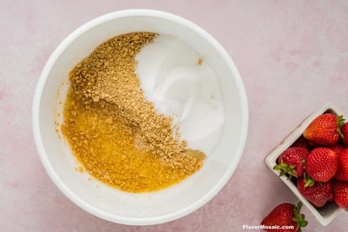 A white bowl with crushed graham crackers, sugar, and melted butter on a pink surface, beside a square dish of fresh strawberries-perfect ingredients for making Strawberry Cheesecake Bars.