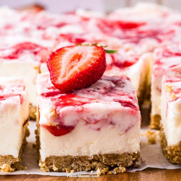 A close-up of creamy Strawberry Cheesecake Bars with a crumbly graham cracker crust, topped with strawberry sauce and a halved fresh strawberry on top.