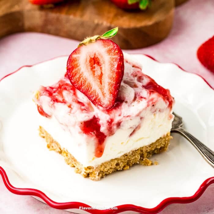 A slice of Strawberry Cheesecake Bars with a graham cracker crust, creamy filling, and strawberry swirl, topped with a halved strawberry, served on a white plate with a red rim and a fork.