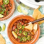 A bowl of pinto bean chili topped with sliced jalapeños and chopped cilantro, served with tortilla chips. Surrounding the bowl are lime wedges, fresh cilantro, another bowl of chili, and a slow cooker.