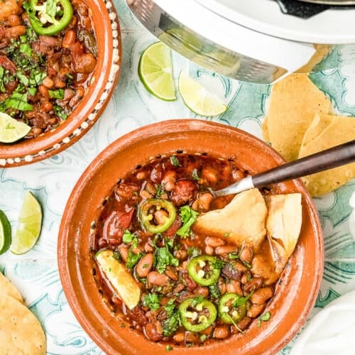A bowl of pinto bean chili topped with sliced jalapeños and chopped cilantro, served with tortilla chips. Surrounding the bowl are lime wedges, fresh cilantro, another bowl of chili, and a slow cooker.