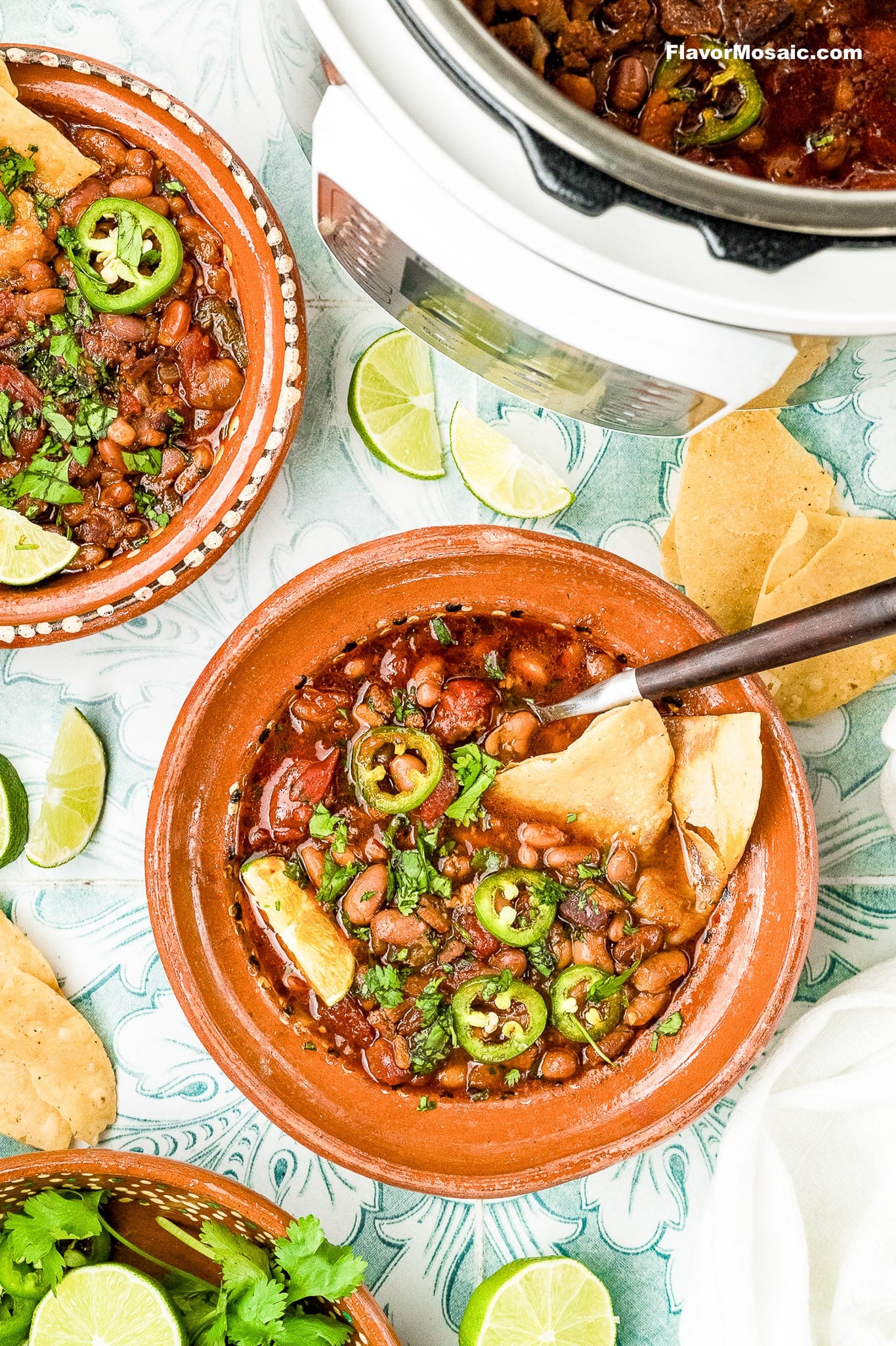A bowl of pinto bean chili topped with sliced jalapeños and chopped cilantro, served with tortilla chips. Surrounding the bowl are lime wedges, fresh cilantro, another bowl of chili, and a slow cooker.