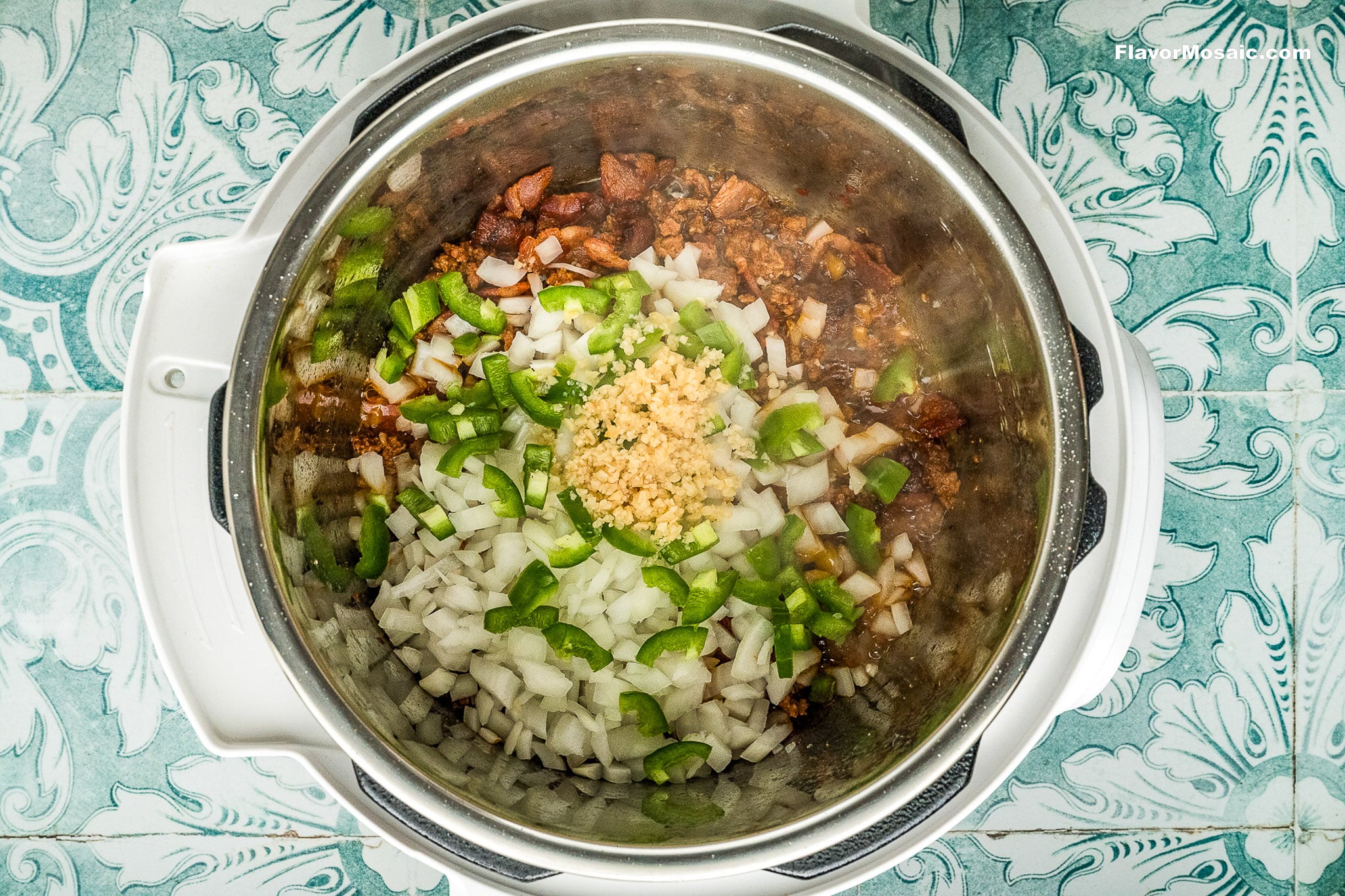 Chopped onions, green bell peppers, and minced garlic sit on browned sausage pieces in an Instant Pot, ready to be mixed and cooked for flavorful Instant Pot Charro Beans (Frijoles Charros). The pot rests on a teal and white patterned surface.