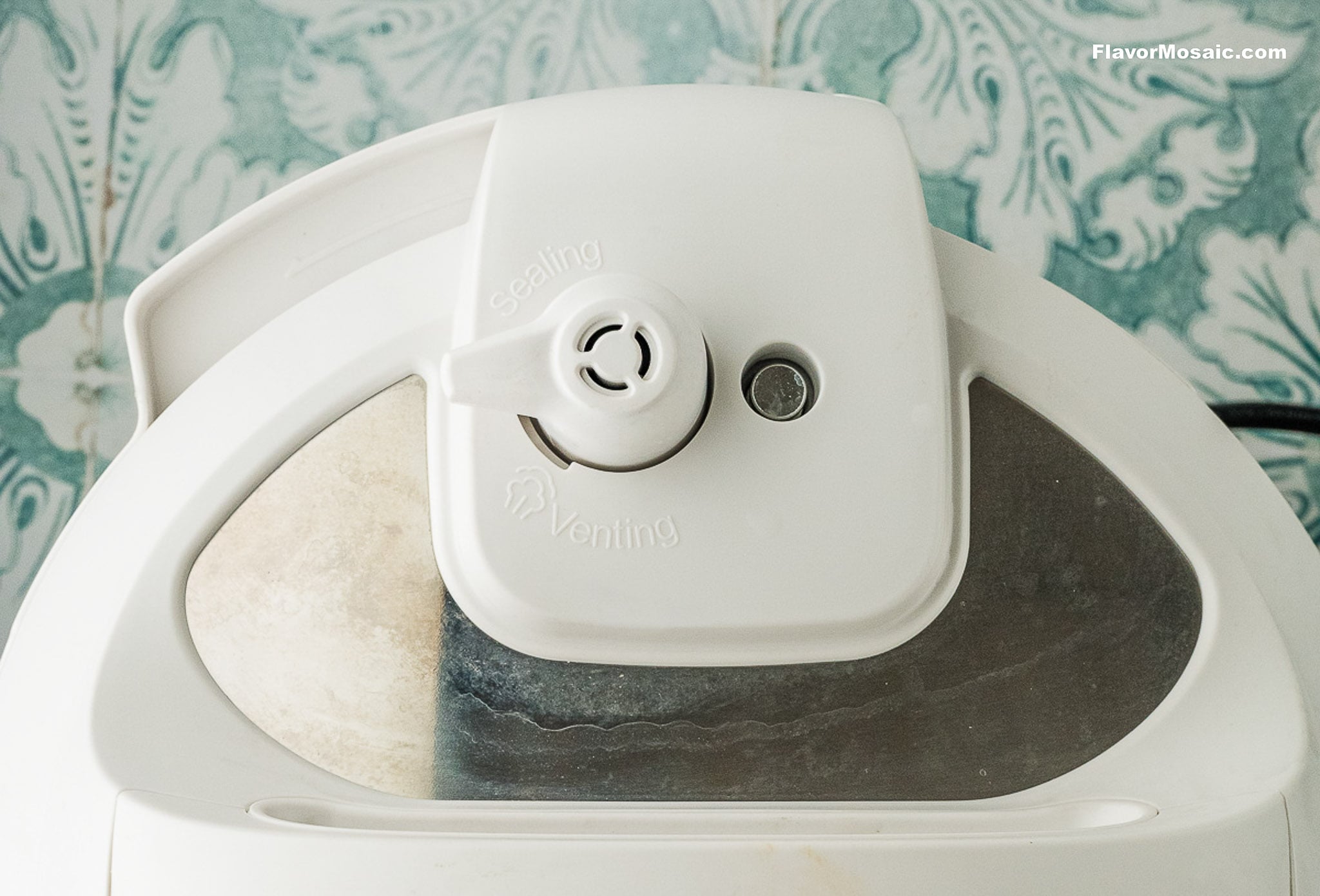 Close-up top view of a white pressure cooker lid showing the sealing and venting steam release valve, perfect for making Instant Pot Charro Beans (Frijoles Charros), with a blue and white patterned background.