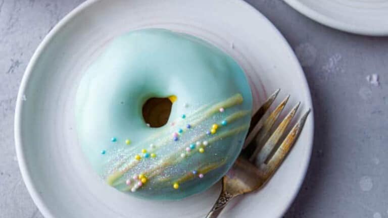 A blue-glazed donut with colorful sprinkles and a drizzle of yellow icing sits on a white plate next to a fork.