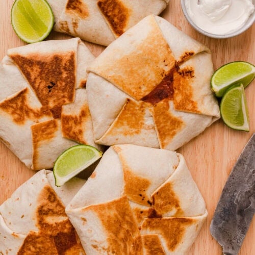 Five folded, grilled tortilla wraps sit on a wooden board with lime wedges and a small dish of sour cream nearby. A knife is placed to the right of the wraps.