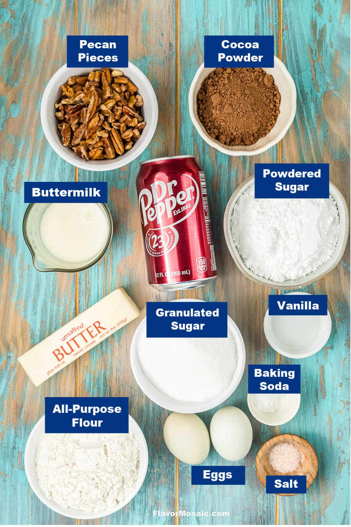A flat lay of baking ingredients labeled: pecan pieces, cocoa powder, buttermilk, powdered sugar, Dr Pepper, granulated sugar, vanilla, butter, flour, eggs, baking soda, and salt on a wooden surface.